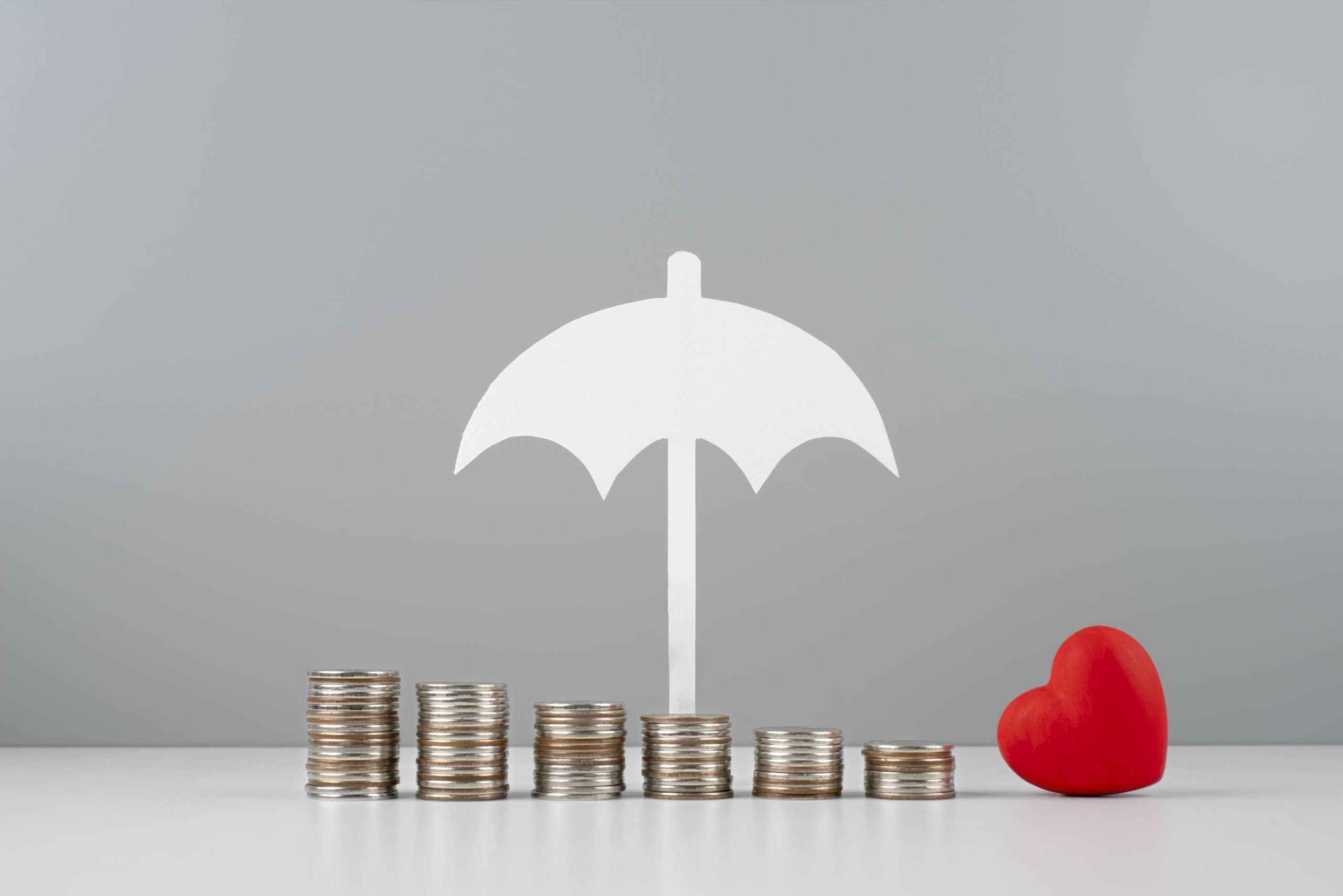 Stacked coins with a white umbrella and a red heart symbolizing insurance savings and protection.