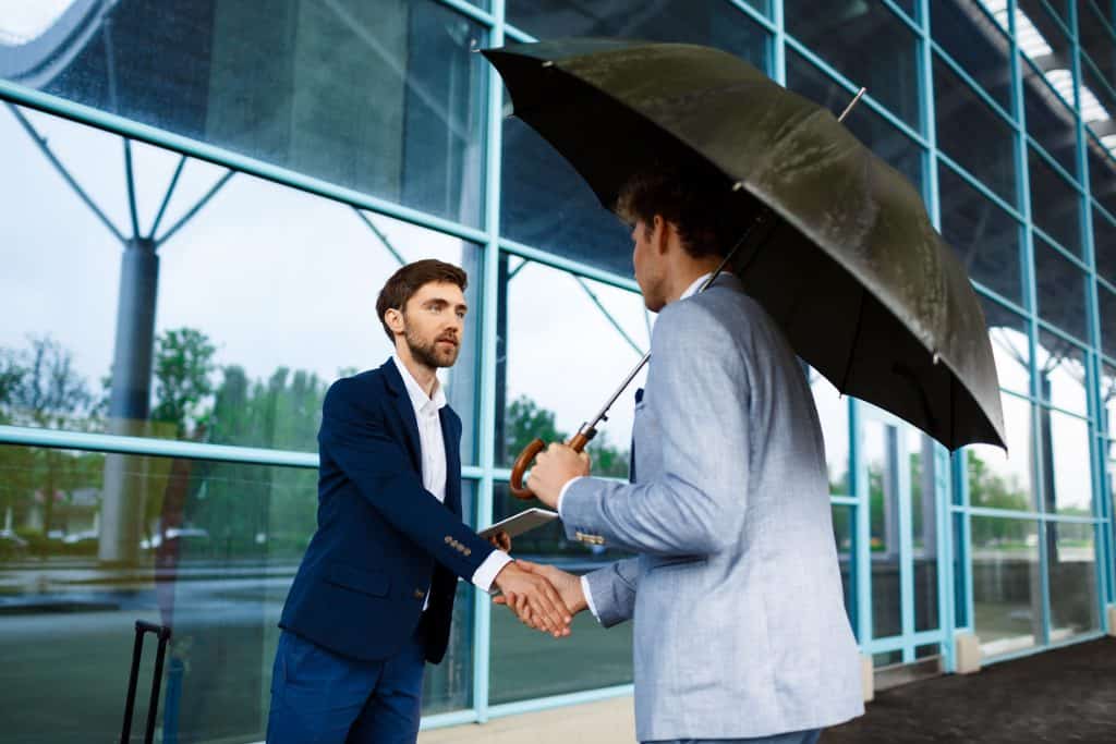 Business professionals shaking hands outside a modern office building under an umbrella.