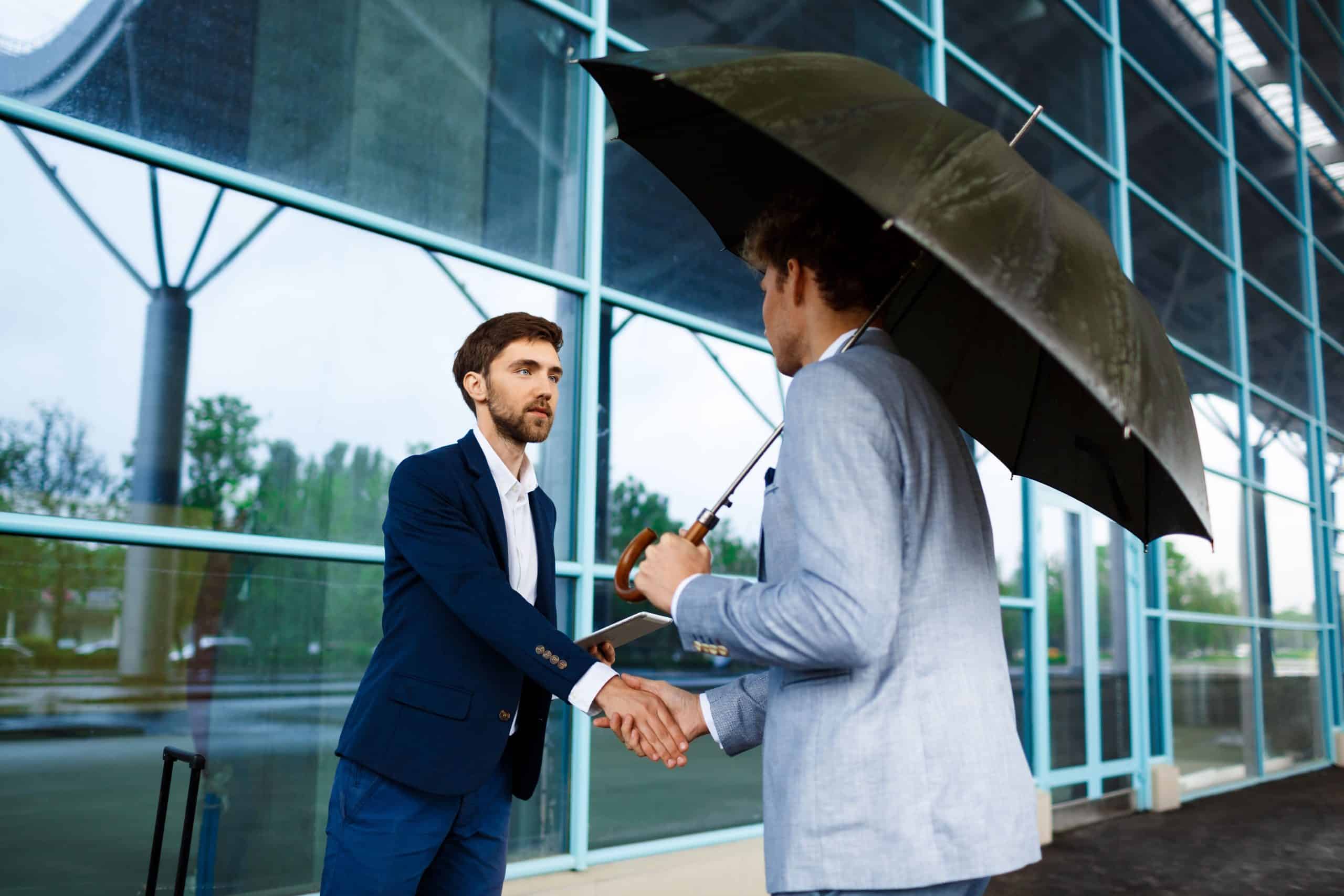 Business professionals shaking hands outside a modern office building under an umbrella.
